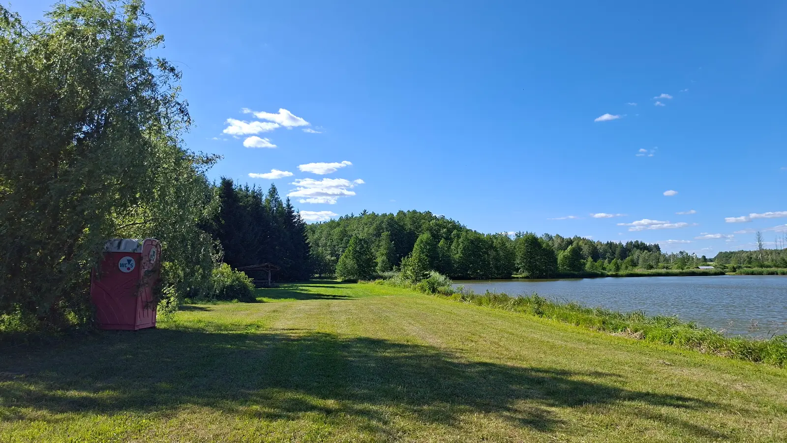 Zdjęcie przedstawia sielski, poziomy krajobraz z jeziorem, utrzymany w formacie panoramicznym, który idealnie nadaje się jako szerokie zdjęcie w tle na stronie internetowej. Po lewej stronie w cieniu dużego, rozłożystego drzewa stoi czerwona przenośna toaleta typu TOI TOI z widocznym logotypem „WC”. Przed nią rozciąga się rozległa, zielona łąka, która prowadzi wzrok widza w stronę spokojnej tafli wody zajmującej prawą część kadru. W oddali, za jeziorem, widać gęsty, mieszany las iglasto-liściasty oraz bezchmurne, jasnoniebieskie niebo z kilkoma delikatnymi obłokami. Kompozycja zdjęcia opiera się na naturalnym kontraście między zacienionym pierwszym planem a słonecznym, otwartym terenem rekreacyjnym nad wodą.